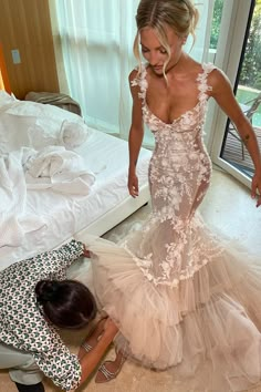 a woman in a wedding dress standing next to another woman on the floor near a bed