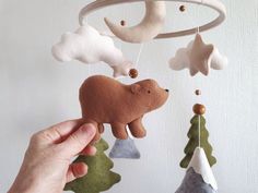 a hand holding a bear mobile in front of a white wall with trees and clouds hanging from it