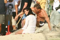 a shirtless man and woman sitting on the sand in front of a group of people