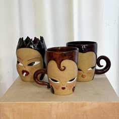 three coffee mugs with faces painted on them are sitting on a wooden table next to a white curtain