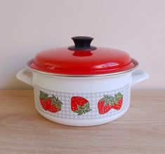 a red and white casserole with strawberries painted on the side sitting on a wooden table