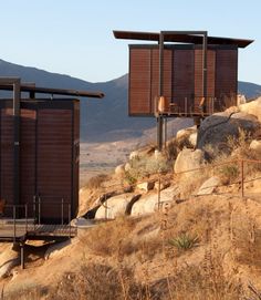 two wooden structures sitting on top of a hill next to dry grass and rocks with mountains in the background