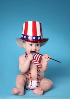 a baby sitting on the ground wearing a patriotic hat and holding an american flag in its mouth