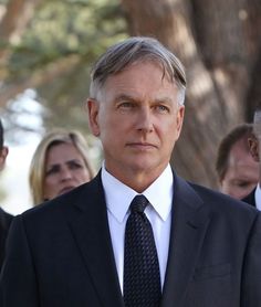 a man wearing a suit and tie standing in front of other people with trees behind him