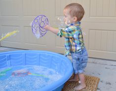 a toddler playing with bubbles in a blue plastic pool while standing next to a garage door