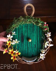 a hand holding a green door decorated with flowers and grass on top of a wooden table