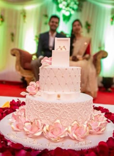 a white wedding cake with pink flowers on the side and a couple sitting behind it