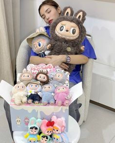 a woman sitting in a chair with several stuffed animals on her lap and one holding a doll