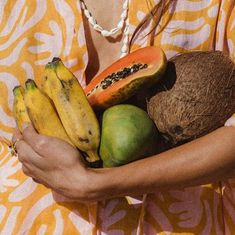 a woman in a yellow and white dress holding a bunch of bananas, watermelon, and kiwi