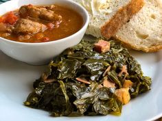 a white plate topped with meat and greens next to a bowl of soup on top of a piece of bread