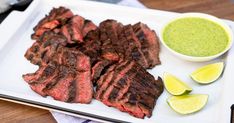 grilled steak with green dipping sauce on a white platter next to sliced limes