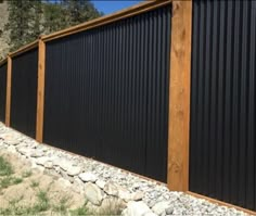 a black metal fence with wooden posts and gravel on the ground next to rocks, grass and trees