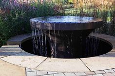 a water fountain in the middle of a garden with purple flowers and grass behind it