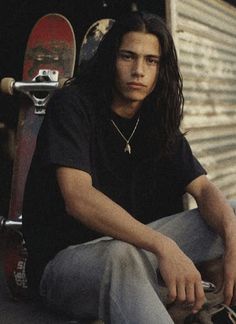 a man with long hair sitting on the ground next to a skateboard and wearing jeans