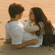 a man and woman sitting on the beach together looking at each other's eyes