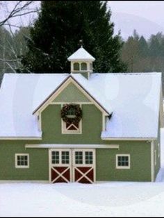 a green barn with a wreath on the door and windows is covered in white snow