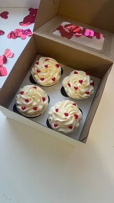 four cupcakes with white frosting and red hearts in a box on a table