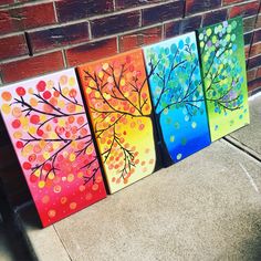 three painted canvass are sitting on the sidewalk next to a brick wall, one has a colorful tree