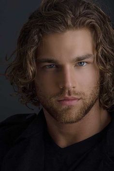 a man with long curly hair and blue eyes looks into the camera while wearing a black shirt