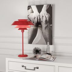 a red lamp sitting on top of a white dresser next to a book and magazine