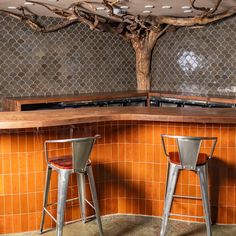 two metal stools sitting in front of an orange tiled bar with a tree on the wall