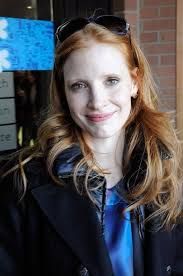 a woman with long red hair wearing a black jacket and blue tie smiling at the camera