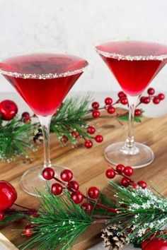 two glasses filled with red liquid on top of a wooden cutting board next to pine cones and berries