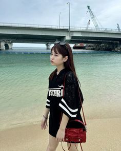 a woman standing on top of a sandy beach next to the ocean with a bridge in the background