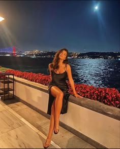 a woman is sitting on a ledge by the water at night with her legs crossed