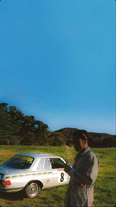 a man standing next to a white car on top of a lush green field under a blue sky