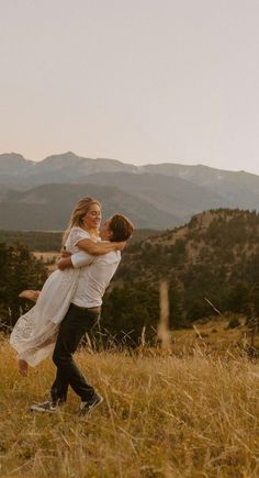 a man and woman are hugging in a field with mountains in the backgroud