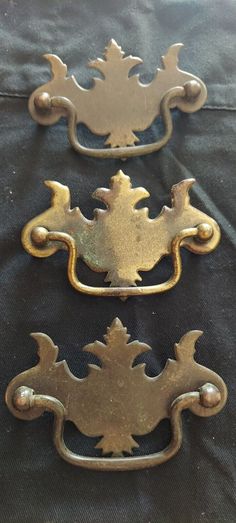 three antique brass drawer pulls on a black fabric background, one has an ornate design and the other has a leaf motif