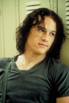 a man with long curly hair wearing a black t - shirt and carrying a green bag