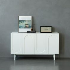 a white cabinet sitting next to a painting on top of a wooden table in front of a gray wall