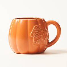 an orange pumpkin shaped coffee mug on a white surface with leaves carved into the cup