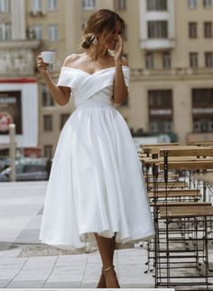 a woman in a white dress holding a cup and talking on her cell phone while walking down the street