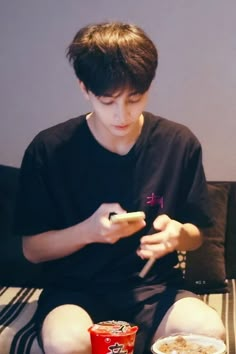 a boy sitting on a couch looking at his cell phone while eating cereal and soda