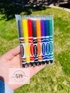 a person holding four different colored crayons in their hand with grass behind them