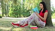 a woman sitting on the ground reading a book next to a tree and stack of books