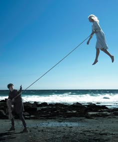 a woman flying a kite on top of a rocky beach next to the ocean with a man