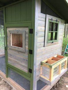 a chicken coop with the door open and two windows on it's side, in front of a green house