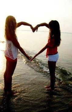 two girls are standing in the water making a heart shape with their hands at sunset