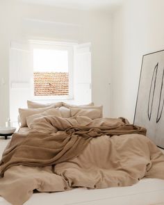 an unmade bed in a white room next to a window with a painting on the wall