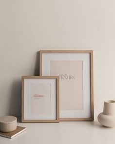 two framed pictures sitting on top of a white table next to a vase and cup