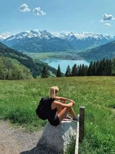 a woman sitting on top of a rock next to a lush green field with mountains in the background