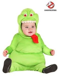 a baby dressed in a green costume sitting on the ground with his mouth open and tongue out