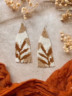 two brown and white beaded earrings sitting on top of an orange cloth next to dried flowers