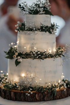 a white wedding cake with greenery and lights on the top is sitting on a tree stump