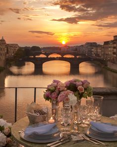 a table is set with plates and silverware in front of a river at sunset