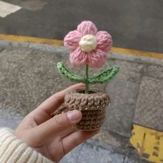 a hand holding a tiny crocheted flower in a pot on the side walk